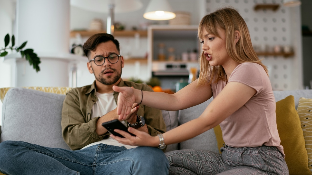 A woman points toward a smartphone screen held by a man sitting on a sofa.