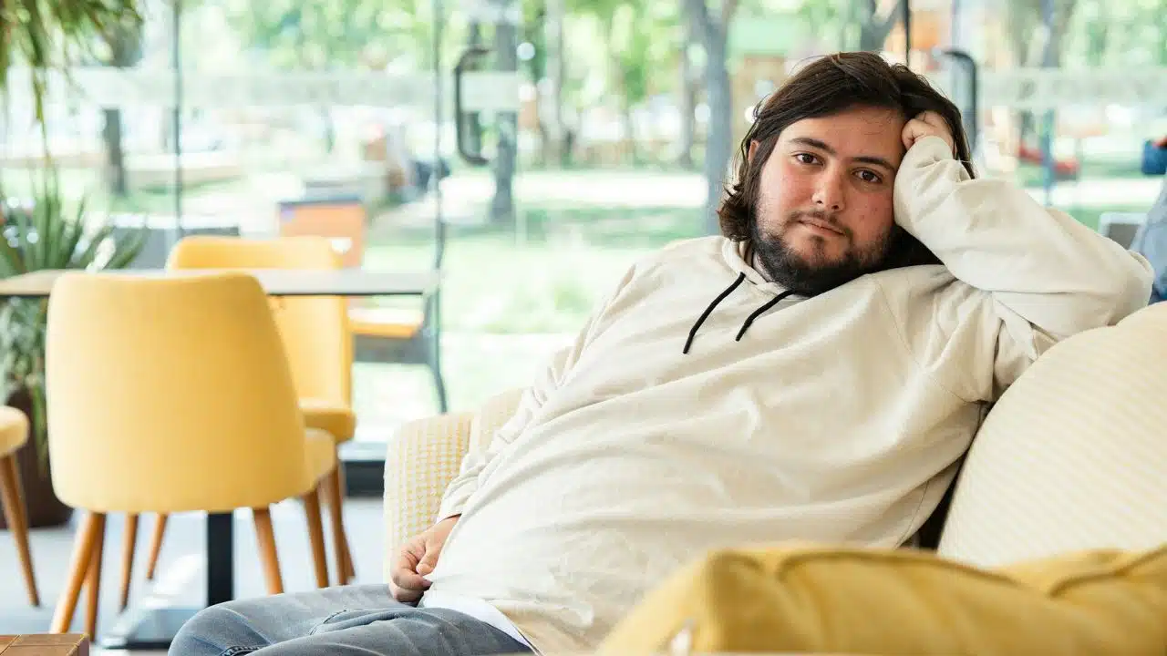 A man in a cream hoodie sits on a sofa, leaning his head on his hand.