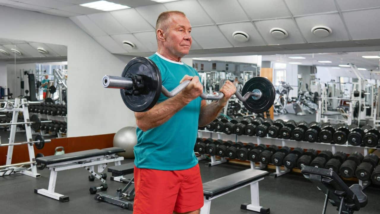 Older man in a green shirt and red shorts lifting a barbell in a gym with weights.