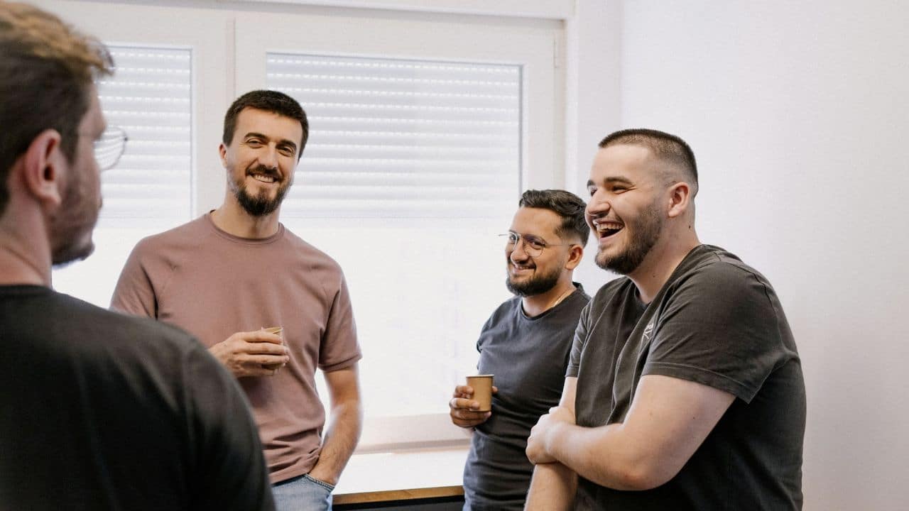 Four men standing indoors, laughing and talking while holding coffee cups.