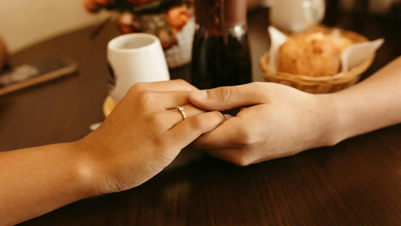 Two people hold hands across a table, with a ring visible on the woman's finger.