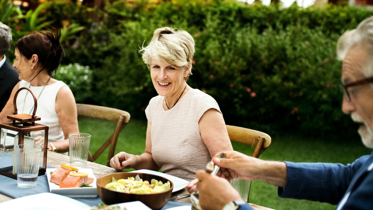 A group of people sits at an outdoor dining table with plates of salmon and salad.