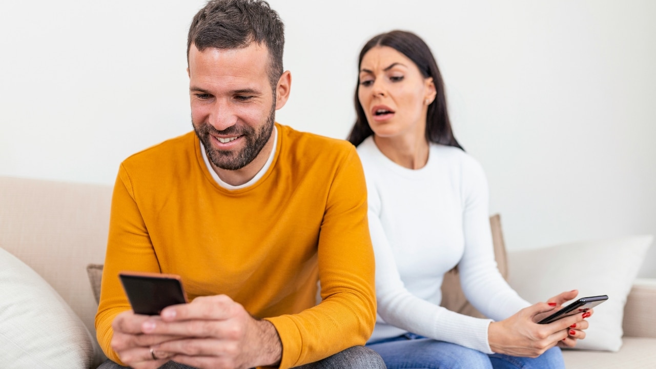 A man smiles at his phone while a woman behind him looks at his screen.