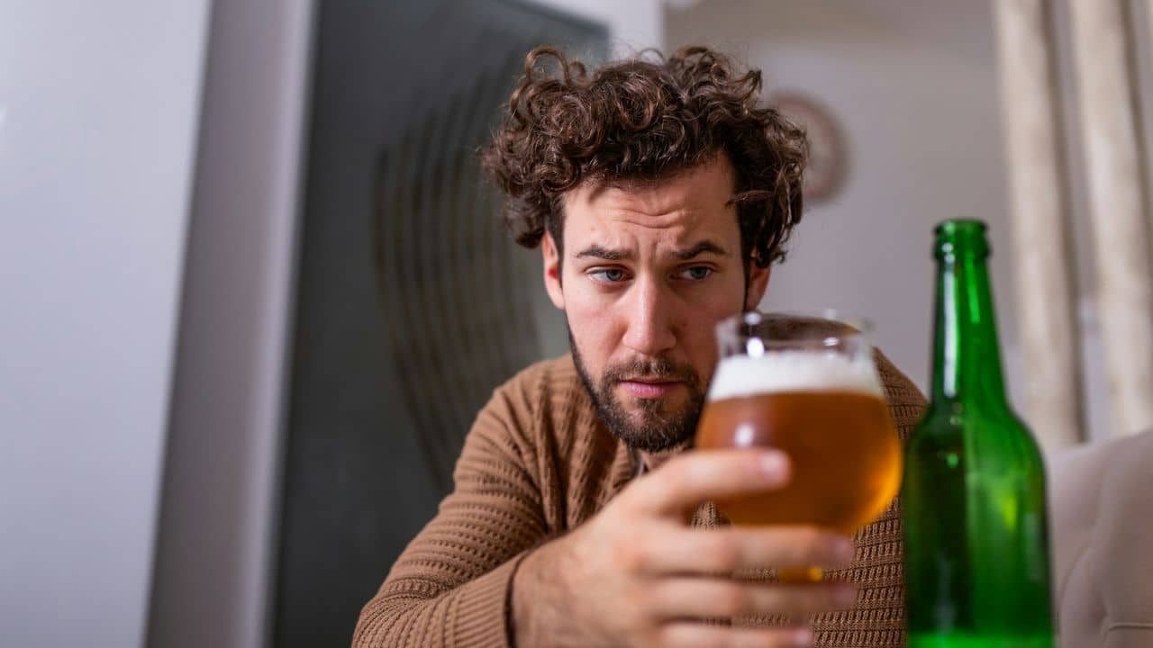 A man with curly hair looks down while holding a glass containing a light liquid.