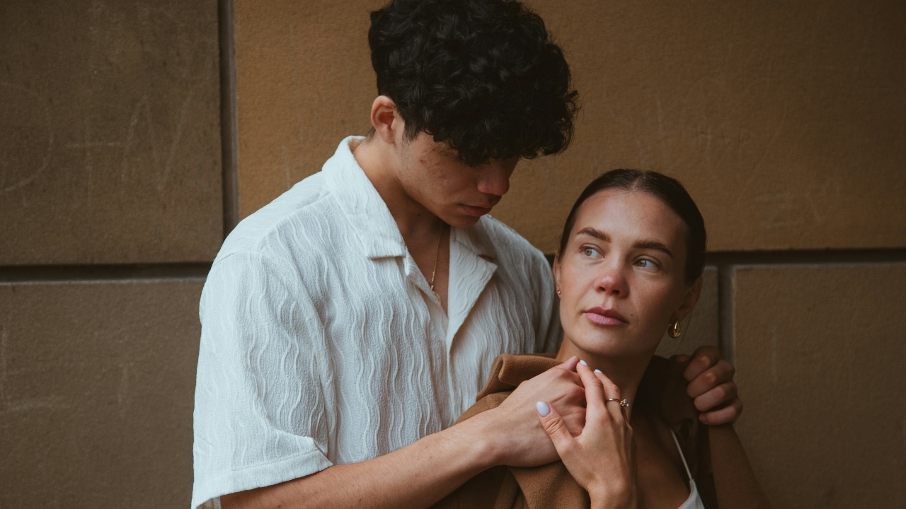 A man stands behind a woman with his hands on her shoulders while she looks away.