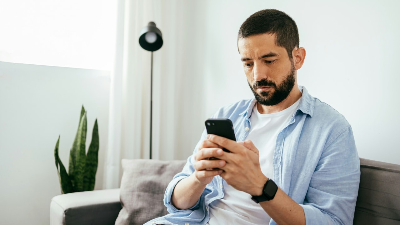 A man with a beard sits on a gray sofa, focused on his smartphone.