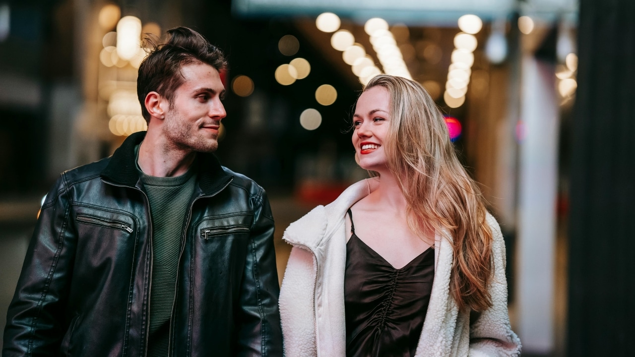 A man and woman walk outdoors at night, looking at each other and smiling.