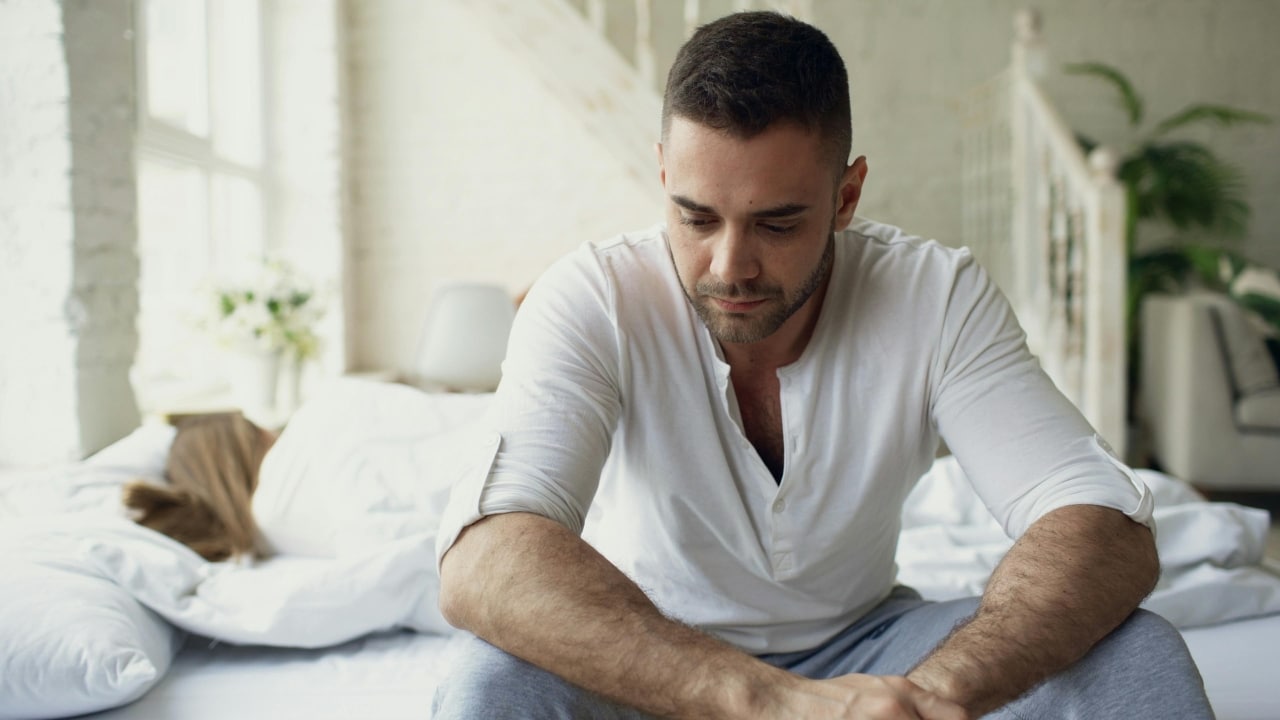 A man sits on the edge of a bed while a woman lies behind him.