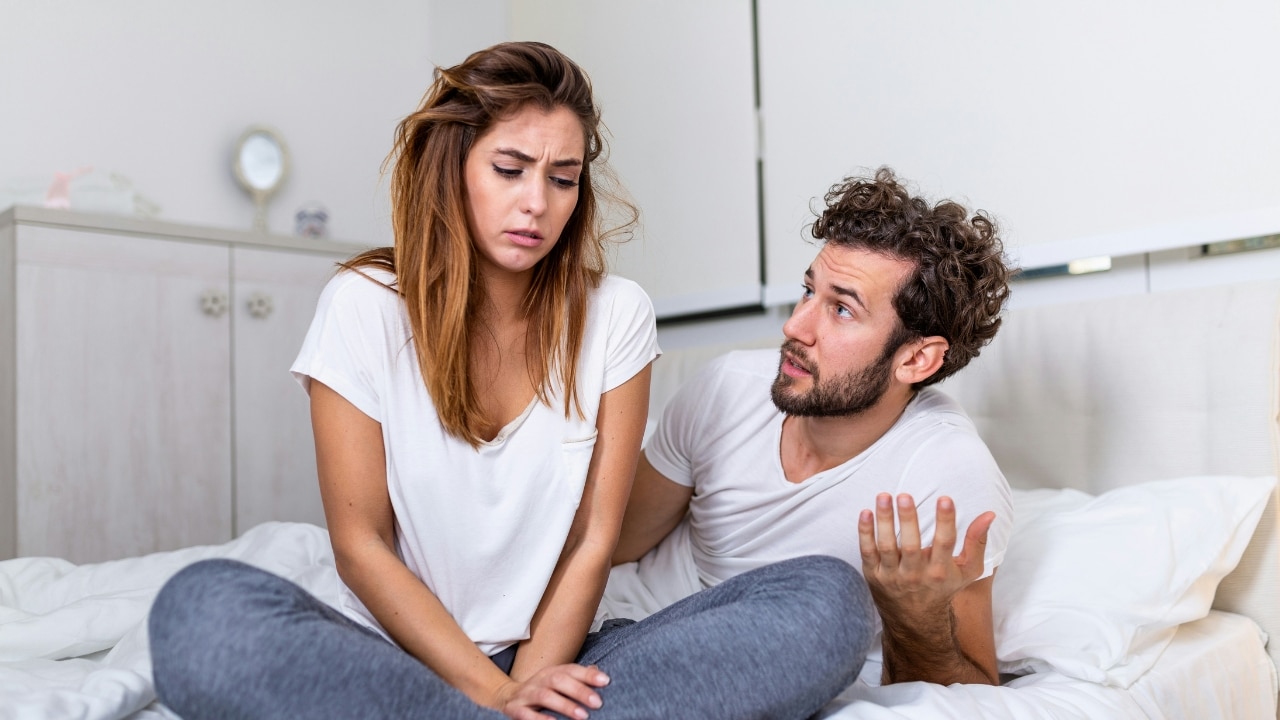 A man and woman sit on a bed while the man gestures with his hand open.