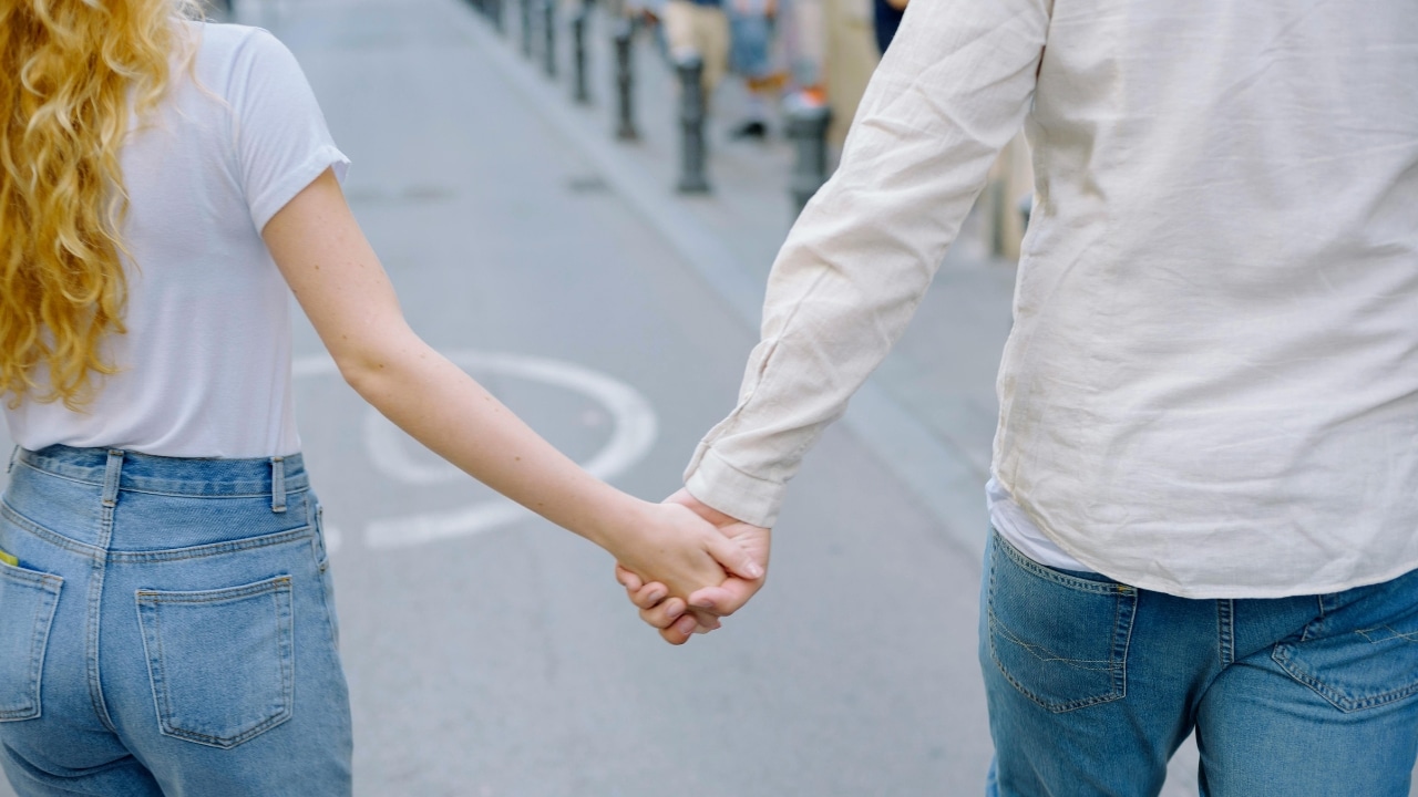 A view from behind of two people holding hands while walking down a paved city street.