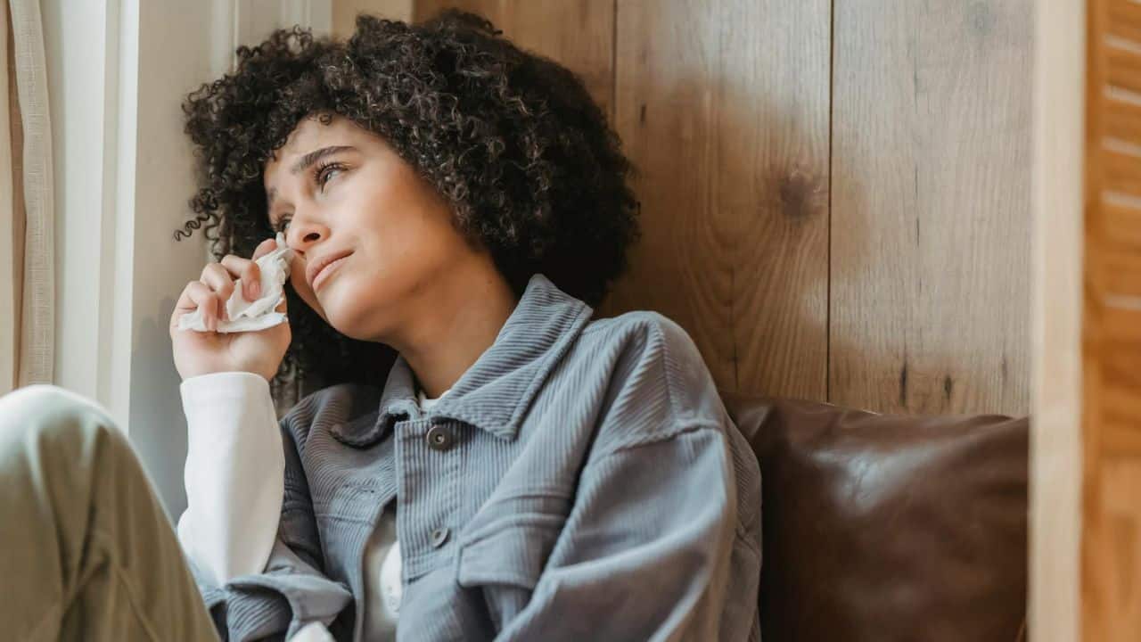 A woman with curly hair sits by a window and wipes tears with a tissue.