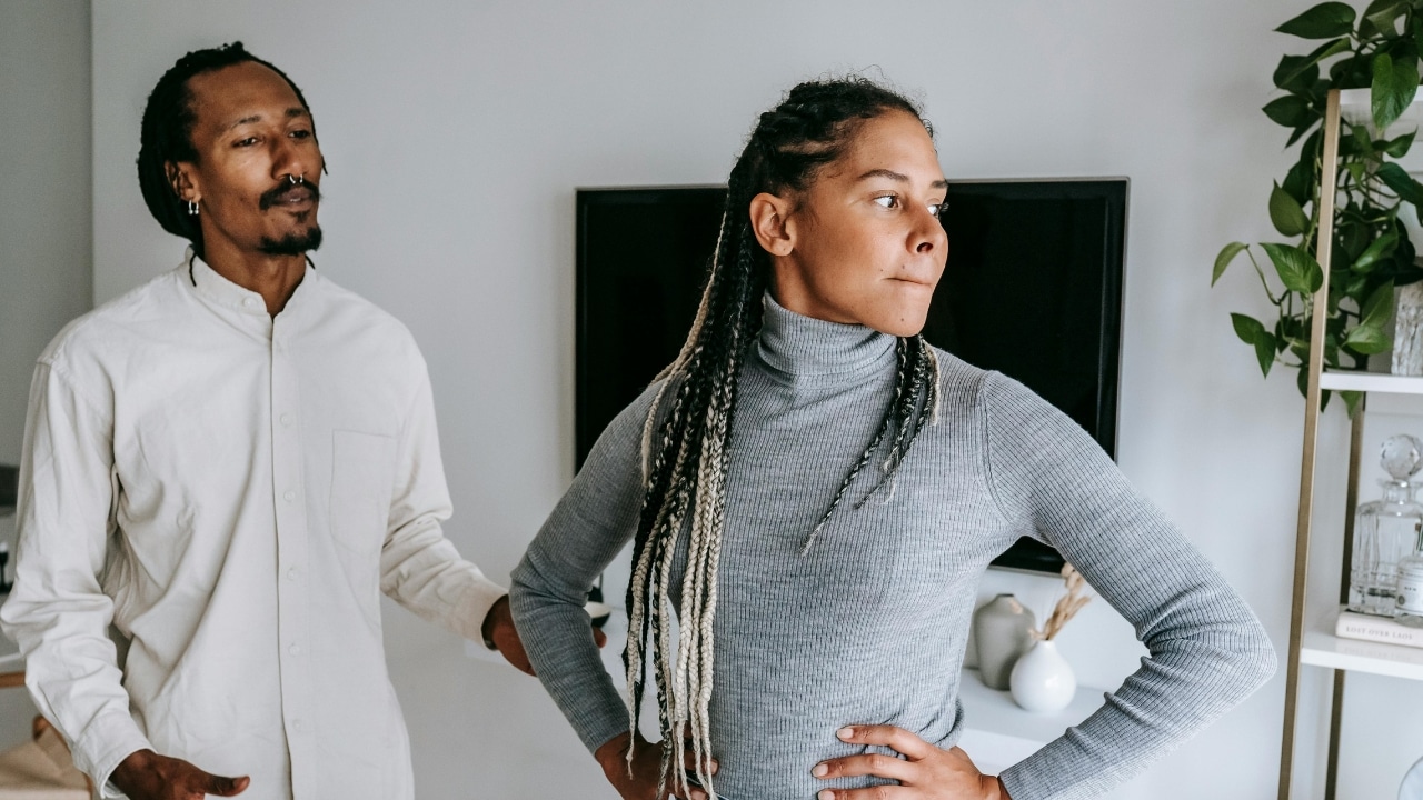 A woman with long braids looks away while a man stands behind her with concern.
