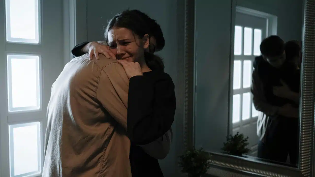 A woman cries while hugging a man in a dark room near a lit door panel.