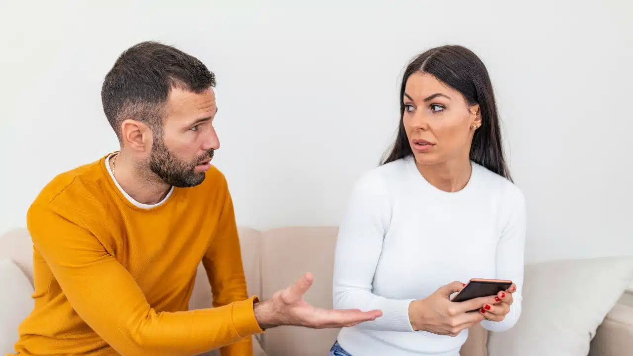 A man gestures with an open hand toward a woman who is looking at a smartphone.