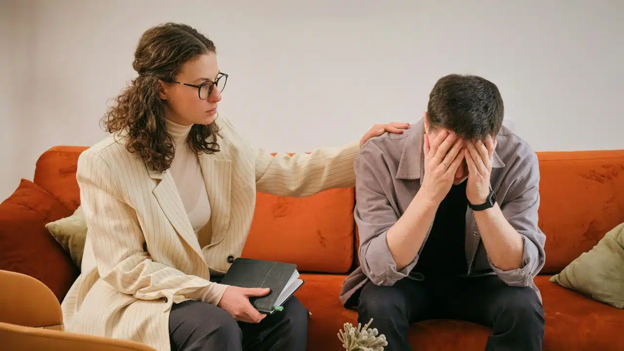 A woman in a blazer places her hand on the shoulder of a distressed man.