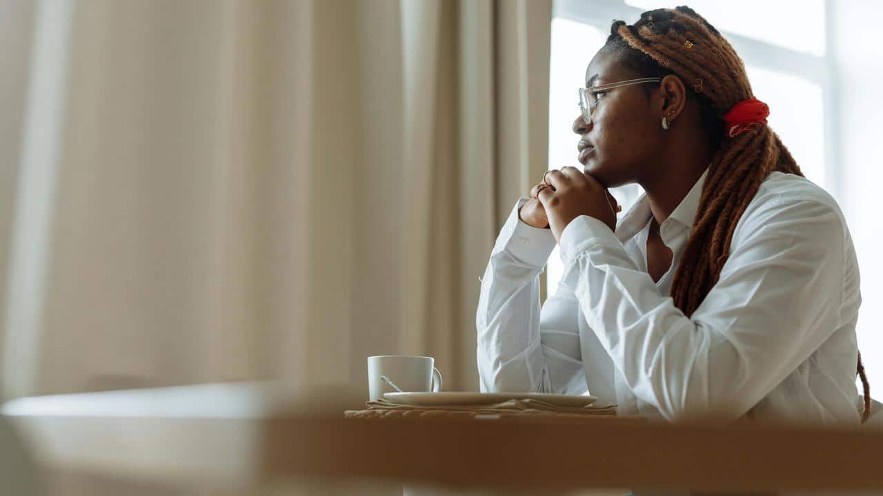 A woman with glasses sits by a window, looking outside thoughtfully.