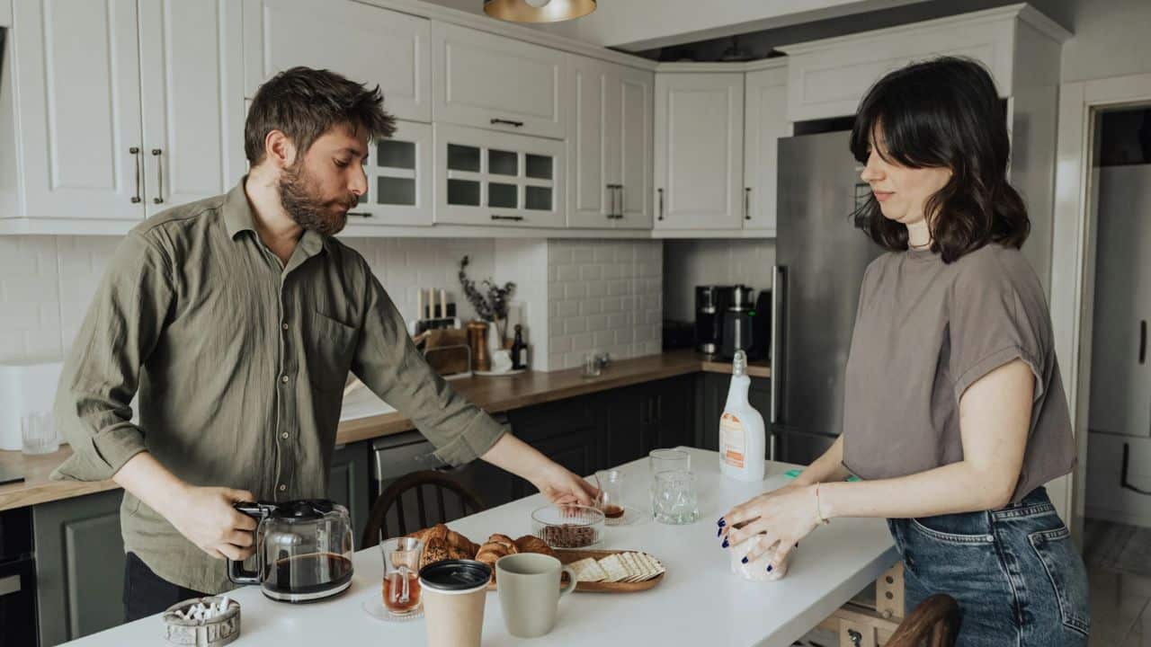 A man and woman stand in a kitchen preparing coffee and food without looking at each other.