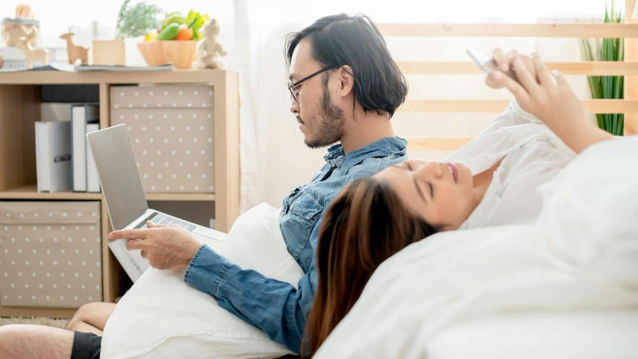 A man uses a laptop while a woman lies beside him looking at a phone.