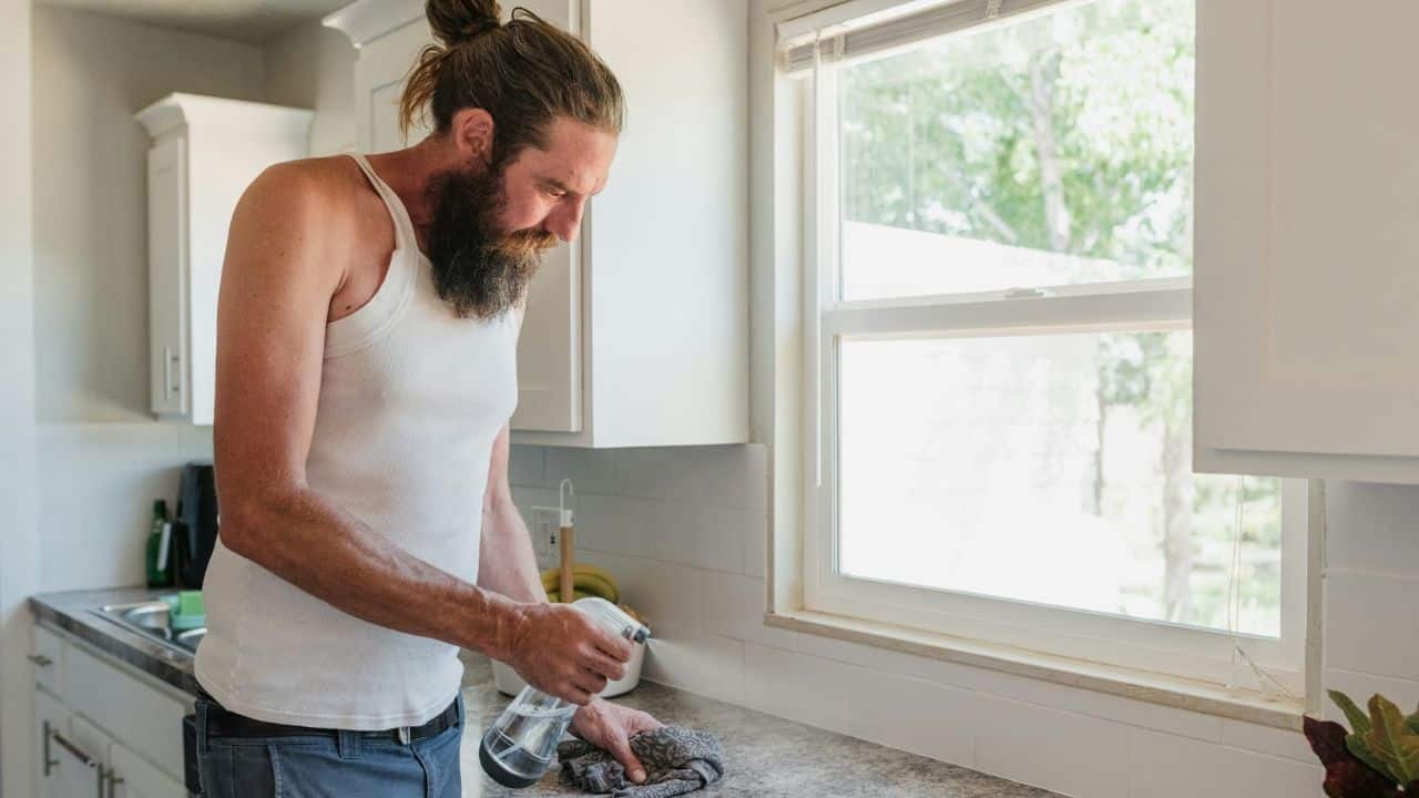 A man with long hair in a bun sprays a kitchen counter while holding a cleaning cloth.