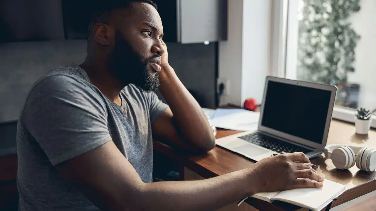 A man sits at a desk with a laptop and notebook, looking thoughtfully out a window.