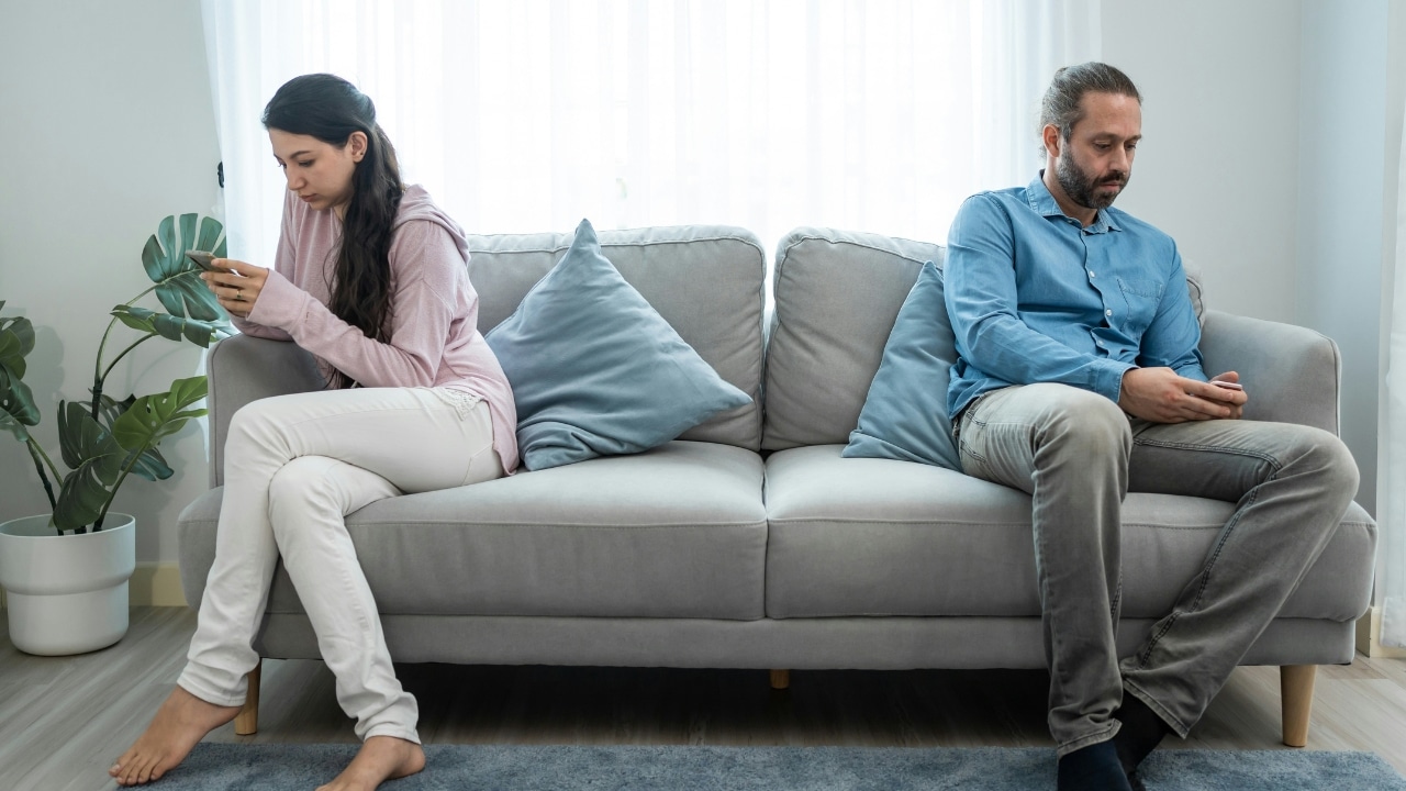 A man and woman sit on opposite ends of a grey sofa, both using their phones.