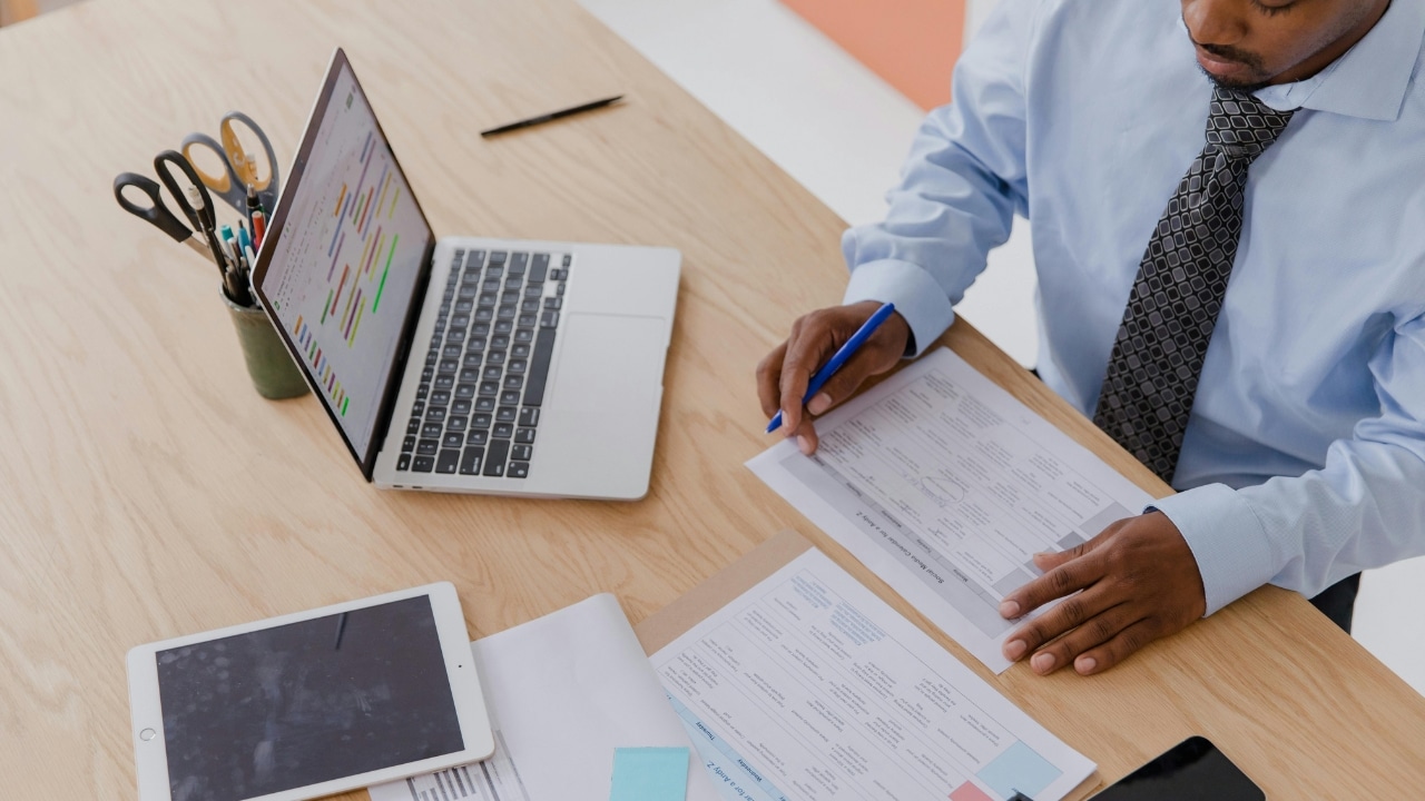 High-angle view of a man in a tie reviewing paperwork at a desk with a laptop.
