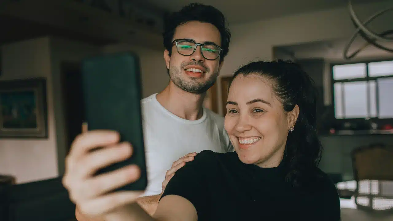 A smiling man and woman pose closely together while holding up a black smartphone for a selfie.