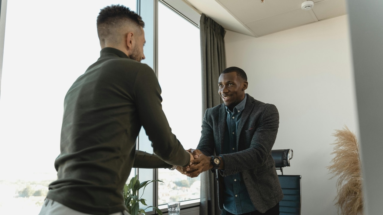 Two men in professional attire shake hands in an office by a large, bright window.