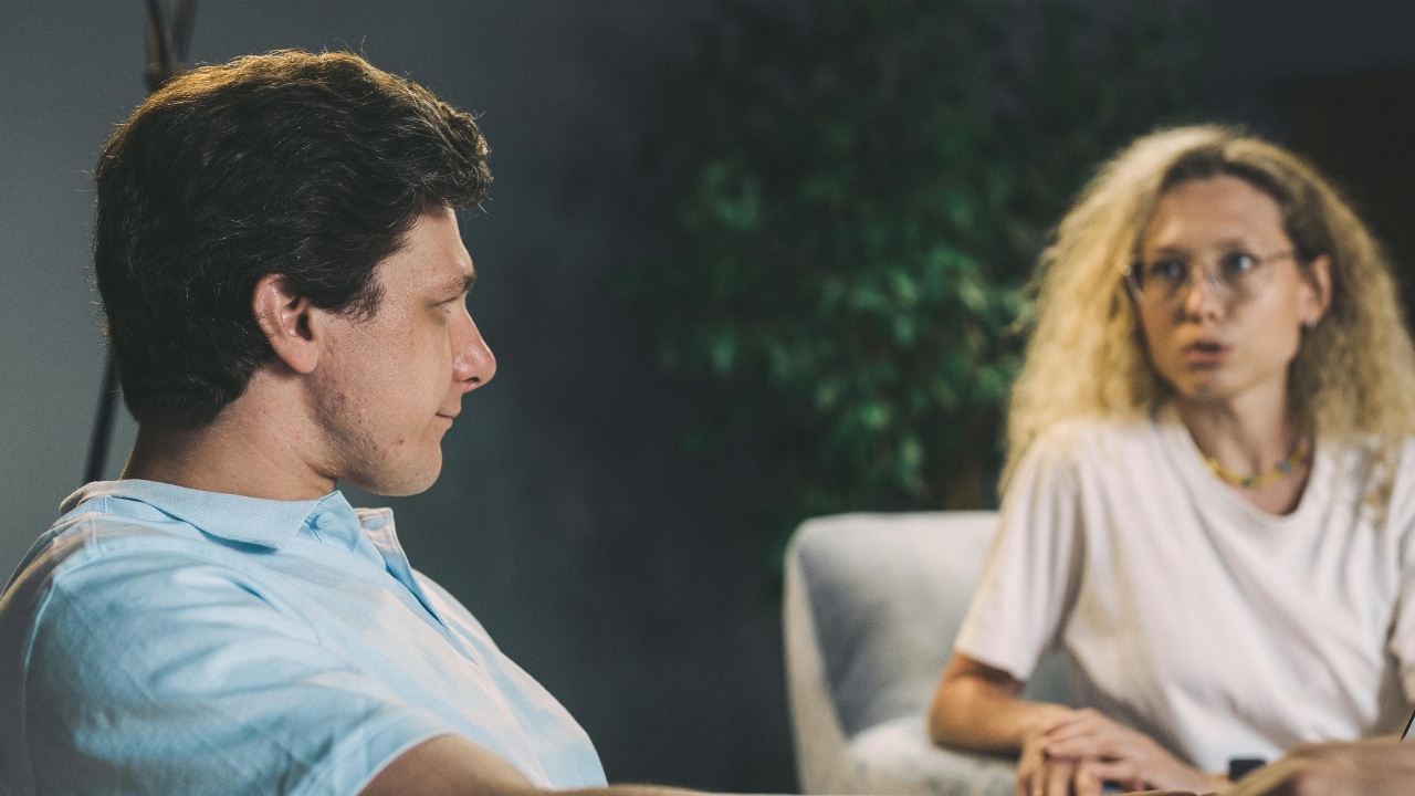 A man in a blue polo shirt looks toward a woman with blonde curly hair.