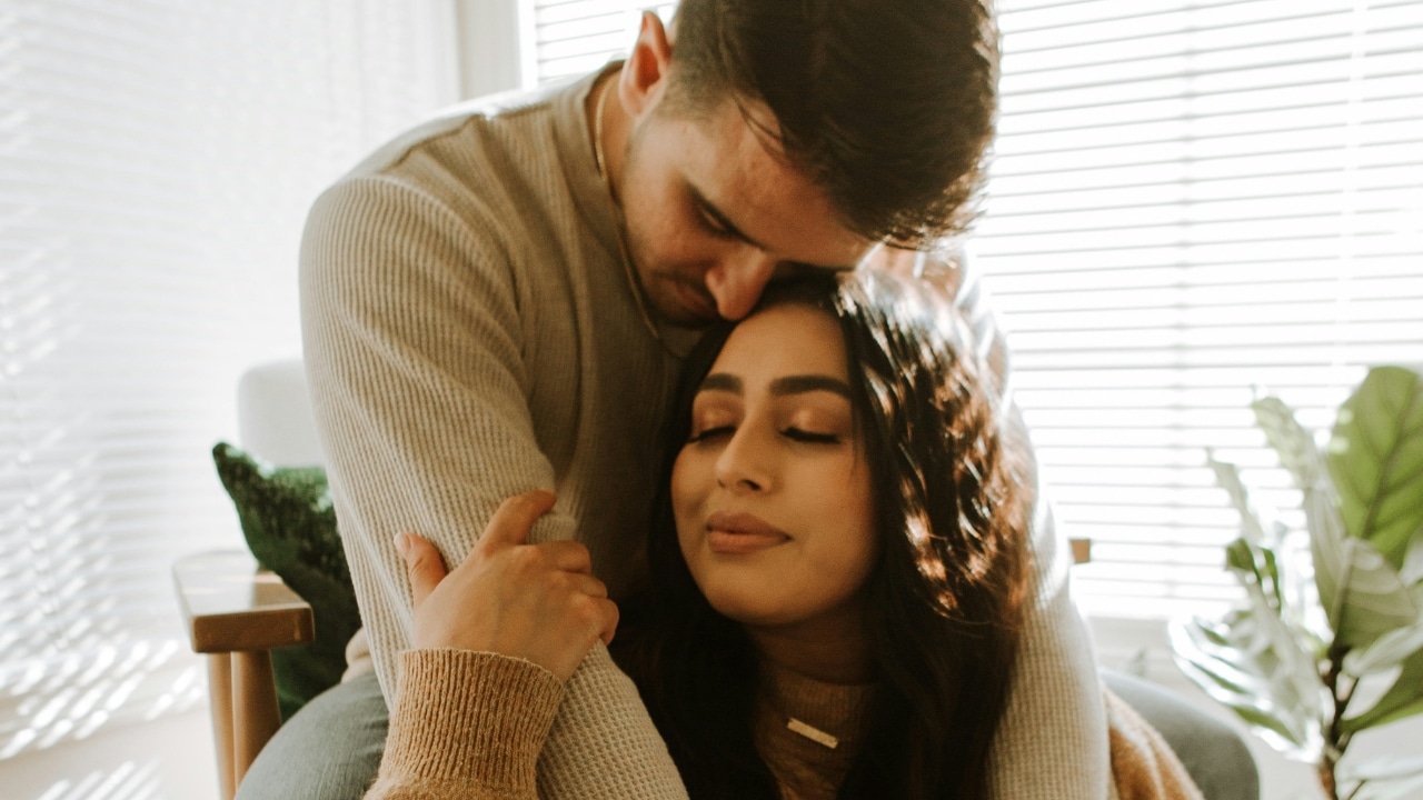 A man hugs a woman from behind while she rests her head back with eyes closed.