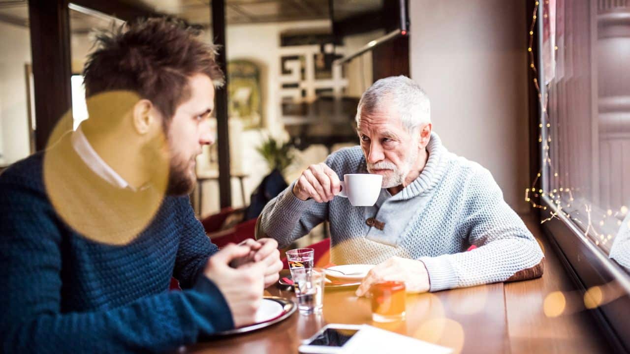 An older man drinks from a cup while sitting across from a younger man.