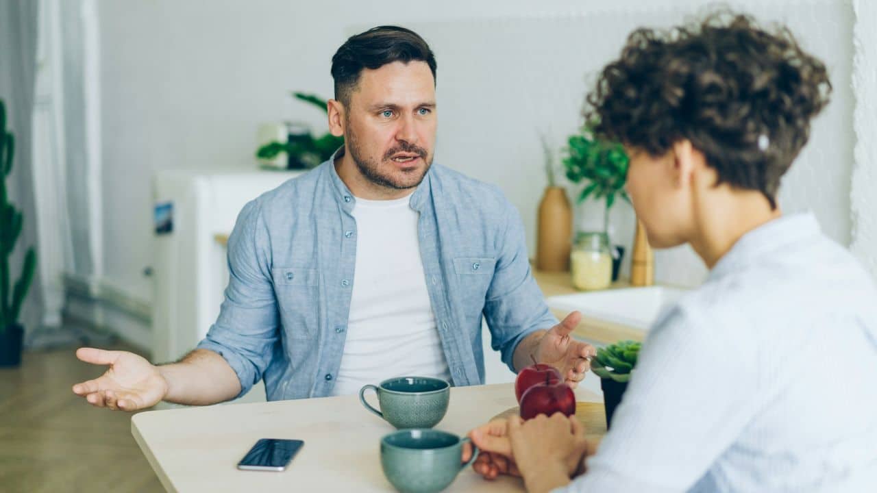 A man gestures while talking to a woman across a kitchen table with mugs and apples.