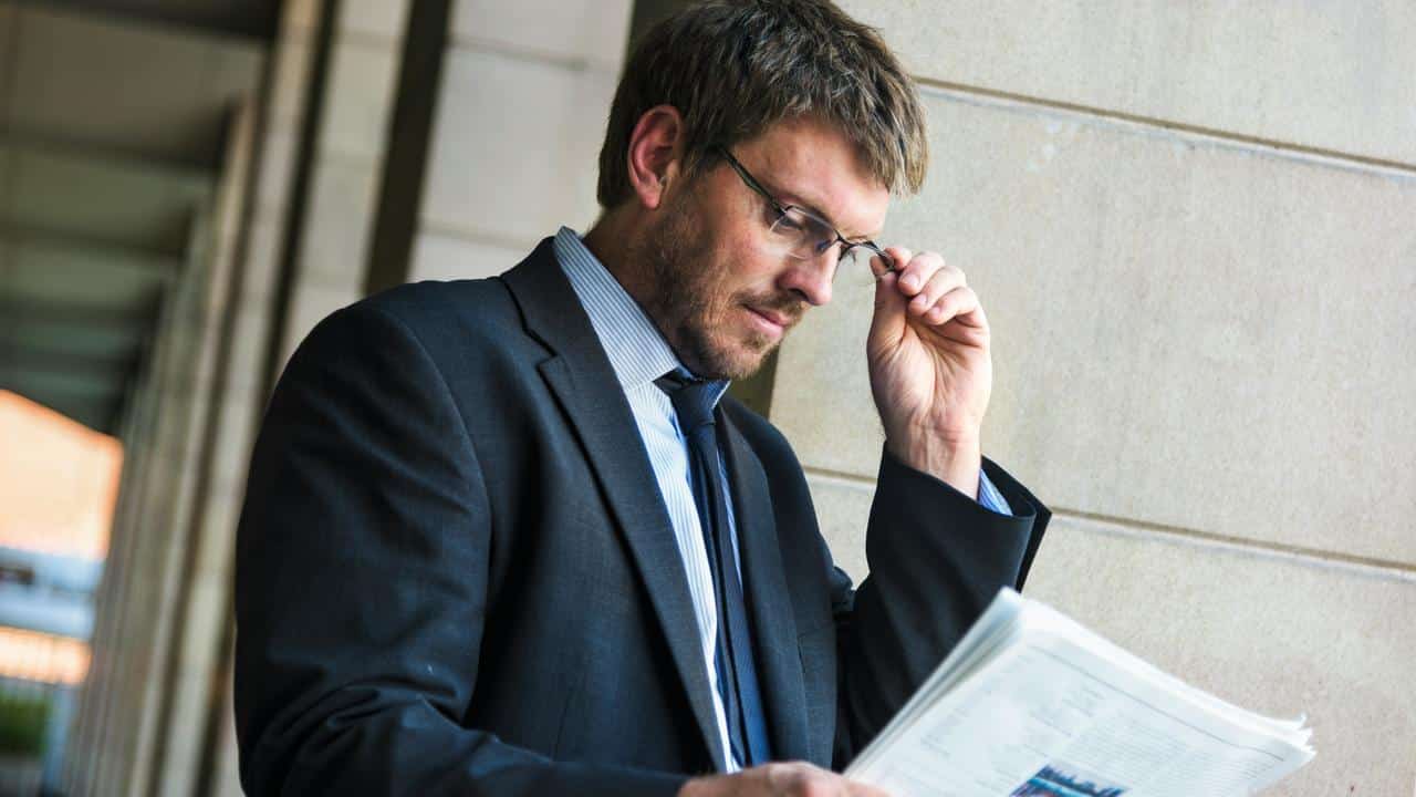 A man in a suit adjusting his glasses while reading a newspaper or document.