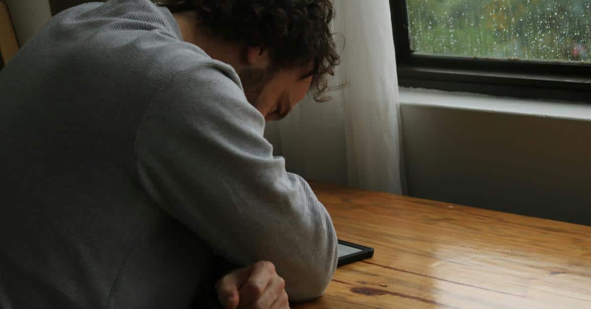 A man resting his head on his arm beside a phone at a wooden table.