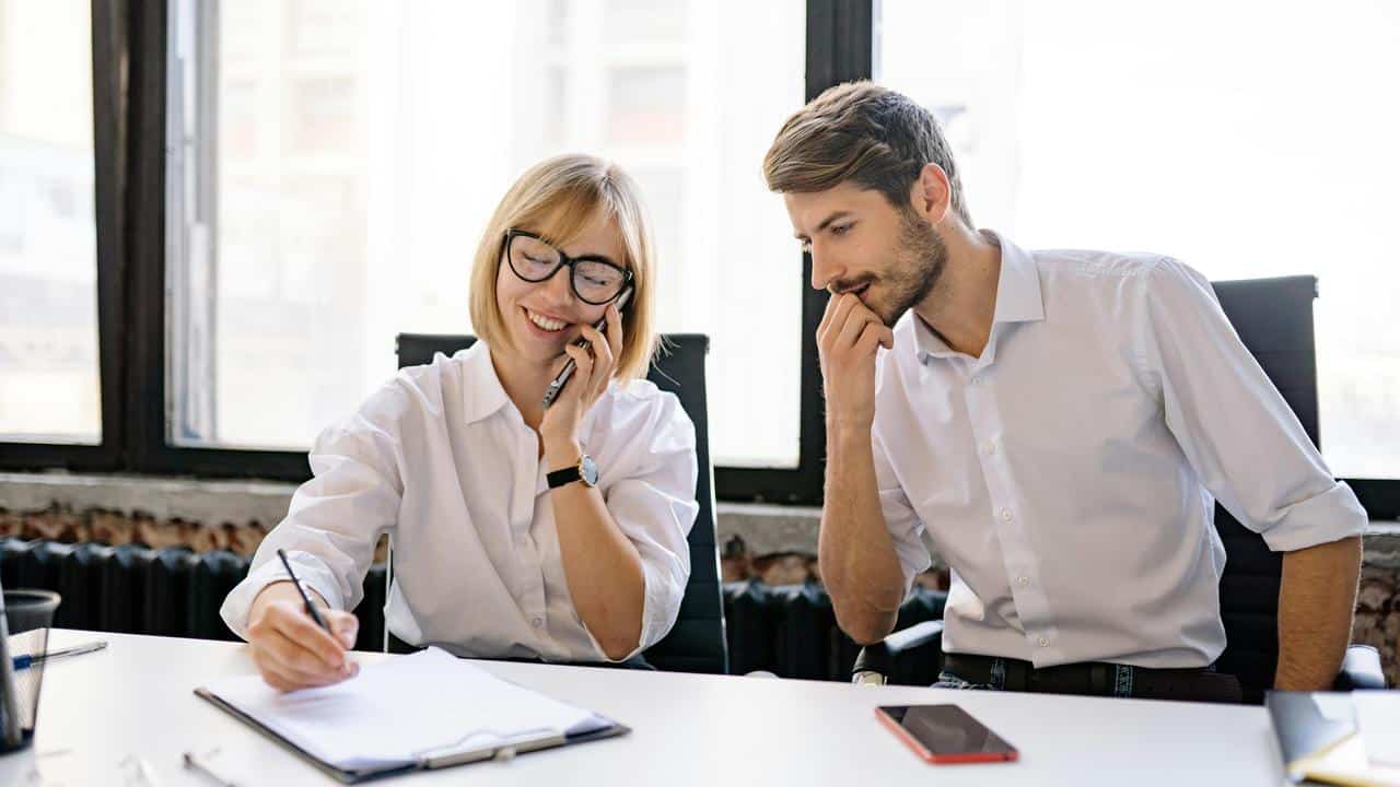 A woman on the phone writing notes while a man watches.