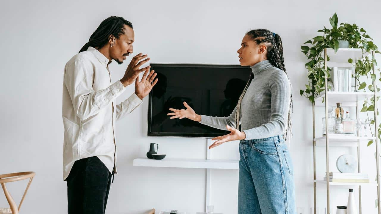 A couple standing indoors, facing each other and arguing with expressive gestures.