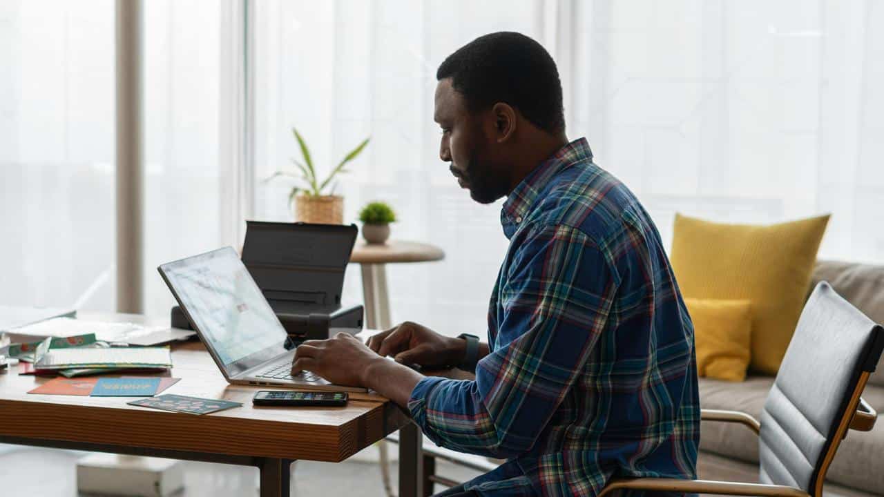 A man sitting at a desk, typing on a laptop in a home office setting.