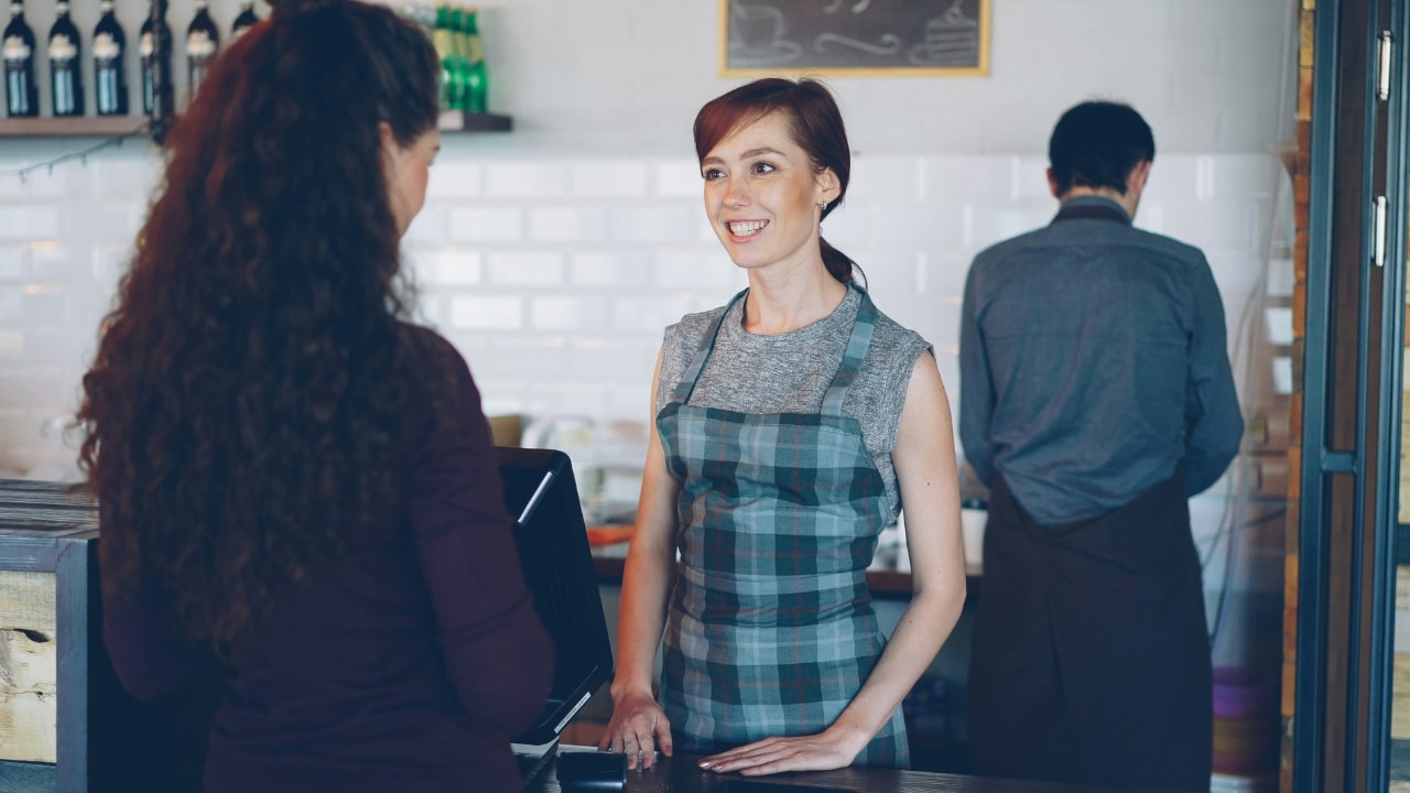 Woman in a plaid apron smiling while standing behind a counter facing a customer.