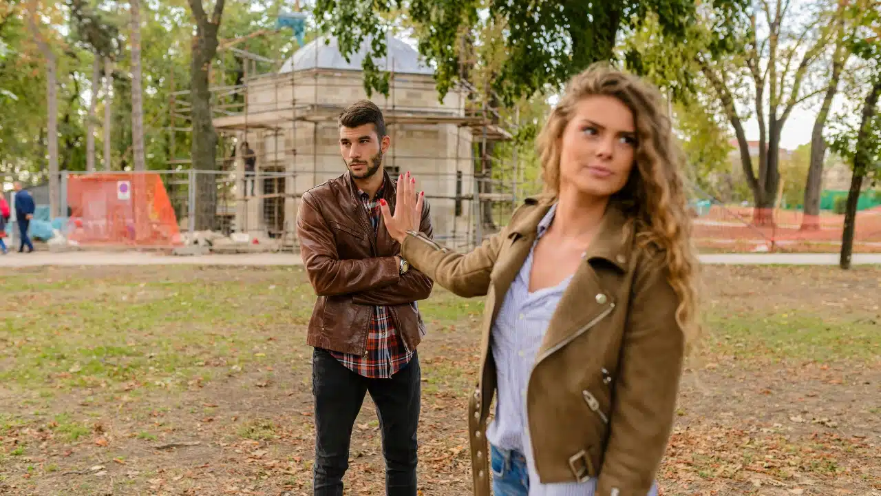 A woman walks away while holding up a hand toward a man standing with crossed arms.