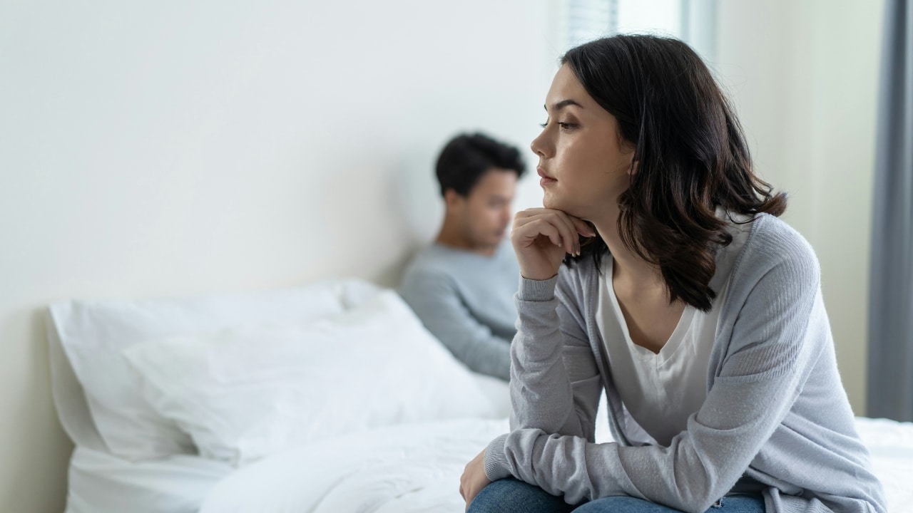 A woman sits on a bed looking away while a man sits blurred behind her.
