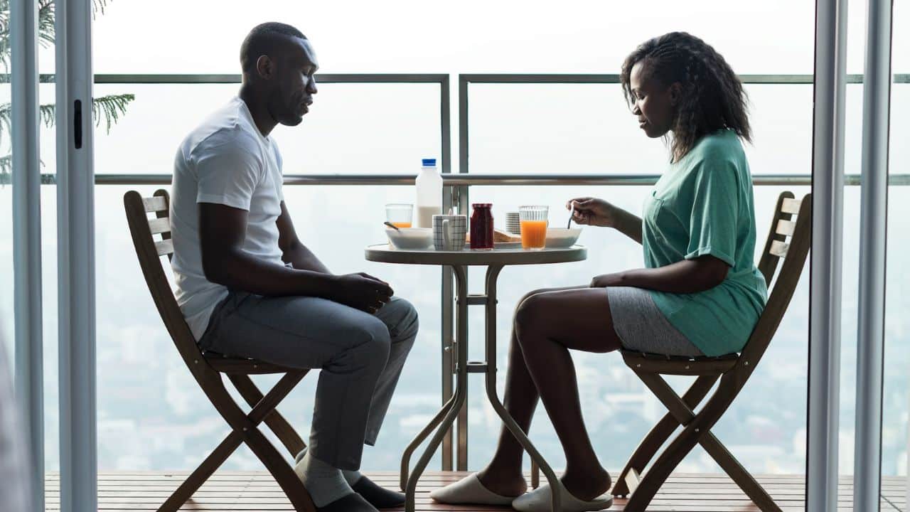 A man and woman sit at a small balcony table eating breakfast in silence.