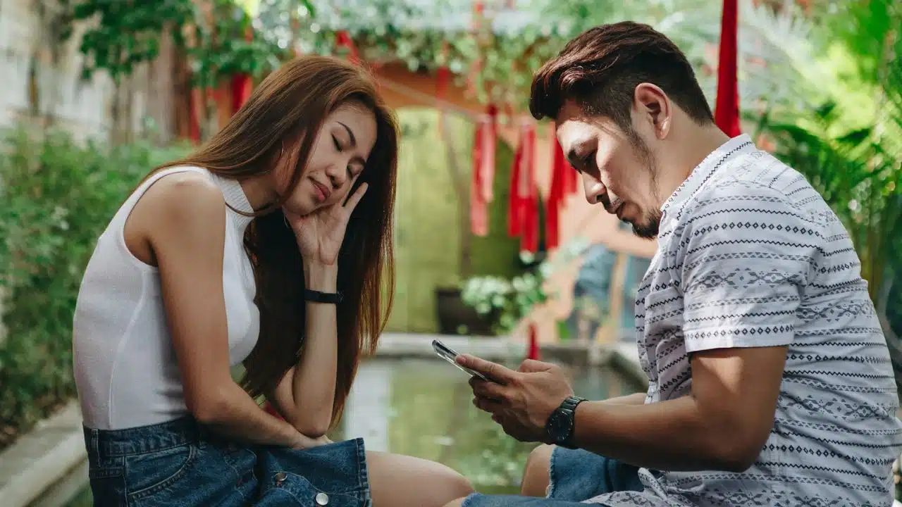 A man looks at a smartphone while a woman sits beside him with her eyes closed.