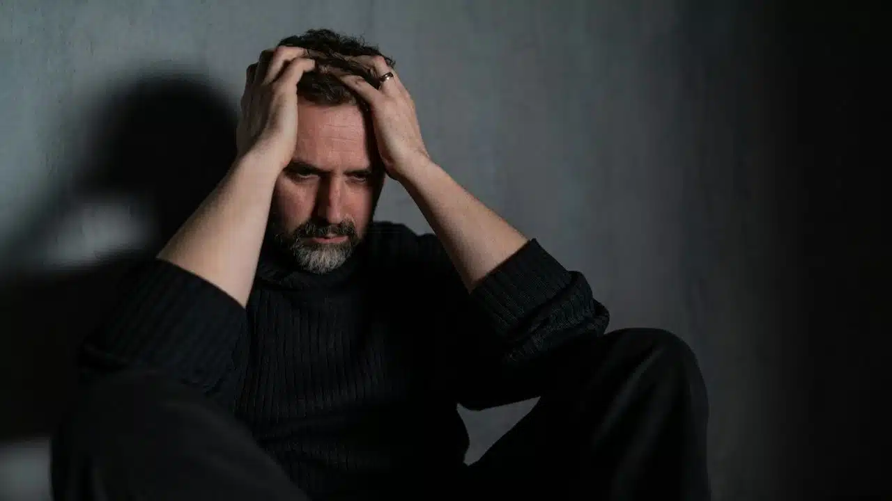 A man in a dark sweater sits against a wall with his hands on his head.