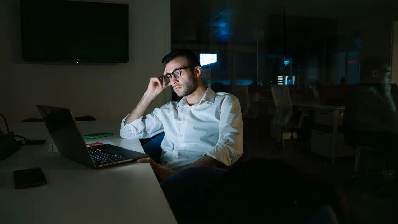 A man in glasses and a white shirt looks tired while working on a laptop in a dimly lit office.