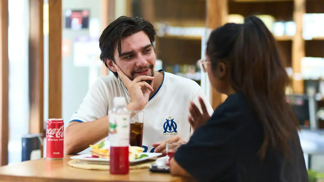 A man in a jersey smiles while listening to a woman across a cafe table.