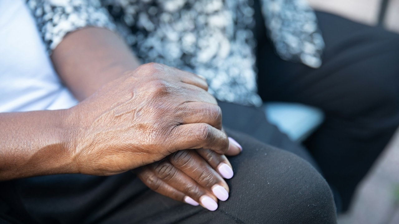 Close-up of two hands clasped on a lap, with one hand featuring light pink nail polish.