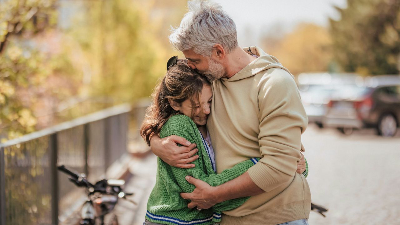A loving father with gray hair hugging his daughter outdoors with a bicycle nearby.