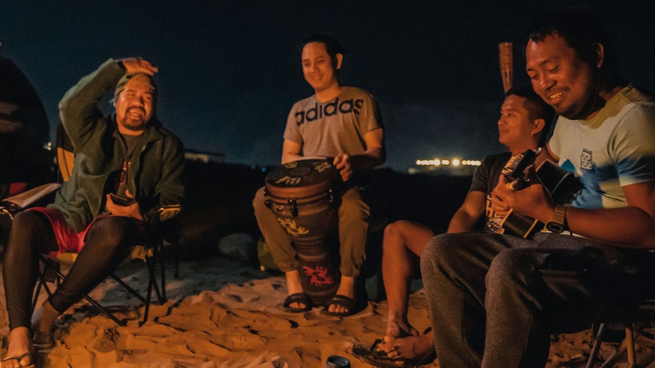Four men sitting on a sandy beach at night, playing music around a campfire.