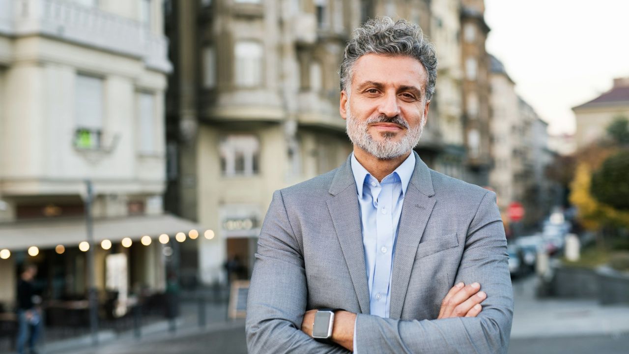 A middle-aged man in a gray suit and blue shirt stands with crossed arms in a city street.