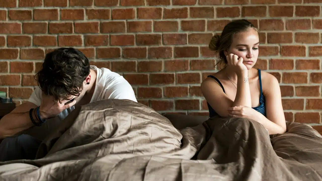 A man and woman sit apart on a bed, both looking down with distressed expressions.